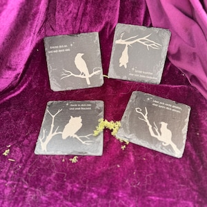 Könnte beinhalten: Vier quadratische Schieferuntersetzer mit geätzten weißen Illustrationen eines Vogels, einer Fledermaus, einer Eule und einer Katze auf Ästen. Jeder Untersetzer hat einen anderen deutschen Satz. Auf einem violetten Samthintergrund.