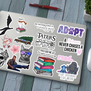 Peut inclure: Un ordinateur portable recouvert de divers autocollants. Les autocollants incluent un dragon noir, un hibou portant des lunettes, un chat avec des lunettes de soleil et du texte qui dit "ADOPT", "A Princess Never Chases a Chicken" et "Stay Different Stay Weird".