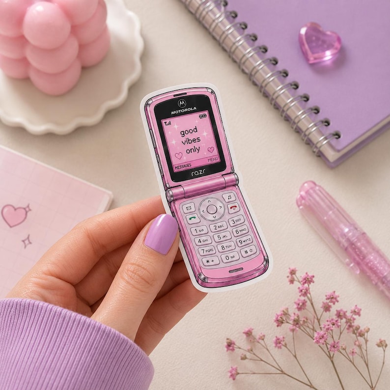 K&ouml;nnte beinhalten: Ein Aufkleber eines rosa Motorola Razr Klapphandys mit einem Bildschirm, der den Text "good vibes only" zusammen mit kleinen Herz- und Sternsymbolen anzeigt. Der Aufkleber zeigt das klassische Tastenfeld, das Motorola-Logo und den Schriftzug razr.