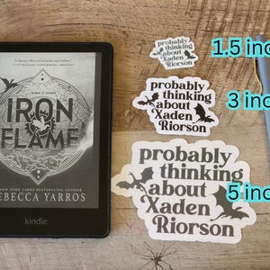 Peut inclure: Un lecteur Kindle affichant le livre "Iron Flame" de Rebecca Yarros. Trois autocollants blancs avec le texte "probably thinking about Xaden Riorson" et des illustrations de chauves-souris sont &agrave; c&ocirc;t&eacute; du lecteur. Un stylo gris est &eacute;galement pr&eacute;sent.