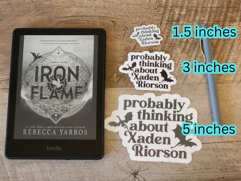 Peut inclure: Un lecteur Kindle affichant le livre "Iron Flame" de Rebecca Yarros. Trois autocollants blancs avec le texte "probably thinking about Xaden Riorson" et des illustrations de chauves-souris sont &agrave; c&ocirc;t&eacute; du lecteur. Un stylo gris est &eacute;galement pr&eacute;sent.