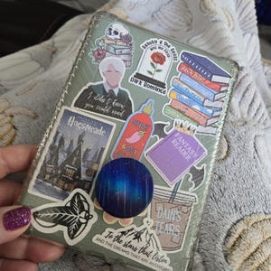 Peut inclure: Une coque de t&eacute;l&eacute;phone transparente orn&eacute;e d'autocollants vari&eacute;s. Les autocollants repr&eacute;sentent un cr&acirc;ne, une rose dans un d&ocirc;me en verre et des piles de livres. Le texte comprend "Dark Romance", "Spicy Book Club", "Fantasy Reader" et "To the stars that listen".