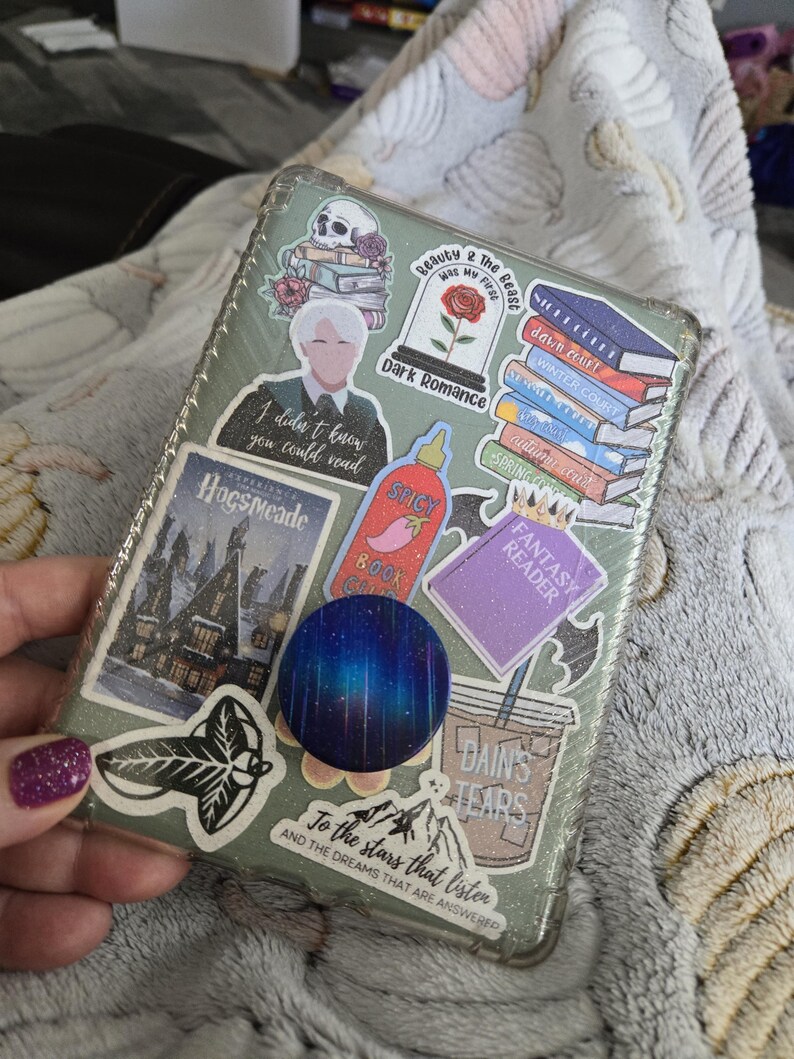 Peut inclure: Une coque de t&eacute;l&eacute;phone transparente orn&eacute;e d'autocollants vari&eacute;s. Les autocollants repr&eacute;sentent un cr&acirc;ne, une rose dans un d&ocirc;me en verre et des piles de livres. Le texte comprend "Dark Romance", "Spicy Book Club", "Fantasy Reader" et "To the stars that listen".