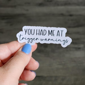 Peut inclure: Un autocollant blanc avec le texte "YOU HAD ME AT trigger warnings" en écriture noire. L'autocollant est tenu par une main avec du vernis à ongles bleu sur un fond en bois flou.