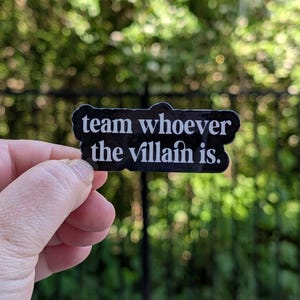 Peut inclure: Un autocollant noir avec du texte blanc qui dit "team whoever the villain is." L'autocollant est tenu devant un fond vert flou. L'autocollant est rectangulaire avec des coins arrondis.