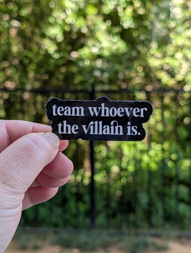 Peut inclure: Un autocollant noir avec du texte blanc qui dit "team whoever the villain is." L'autocollant est tenu devant un fond vert flou. L'autocollant est rectangulaire avec des coins arrondis.