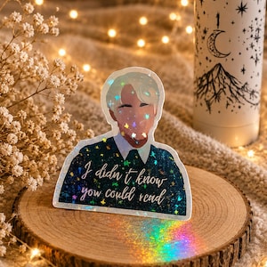 May include: A holographic sticker featuring a stylized illustration of a person with the text "I didn't know you could read." The sticker is placed on a wooden slice, with a white water bottle with a mountain and moon design in the background.