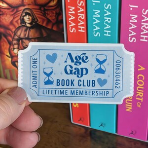 May include: A light blue ticket-shaped sticker with the text "Age Gap Book Club Lifetime Membership." The sticker has a serrated edge and features blue hearts and hourglasses. The number "00630462" is printed on the right side.