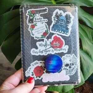 Peut inclure: Une coque de t&eacute;l&eacute;phone transparente avec un fond noir paillet&eacute;, orn&eacute;e de divers autocollants. Les autocollants incluent un pistolet avec le texte "Zade Meadows", des roses, une t&ecirc;te de mort et les mots "Meadows, baby". Un pop socket bleu et violet est attach&eacute;.
