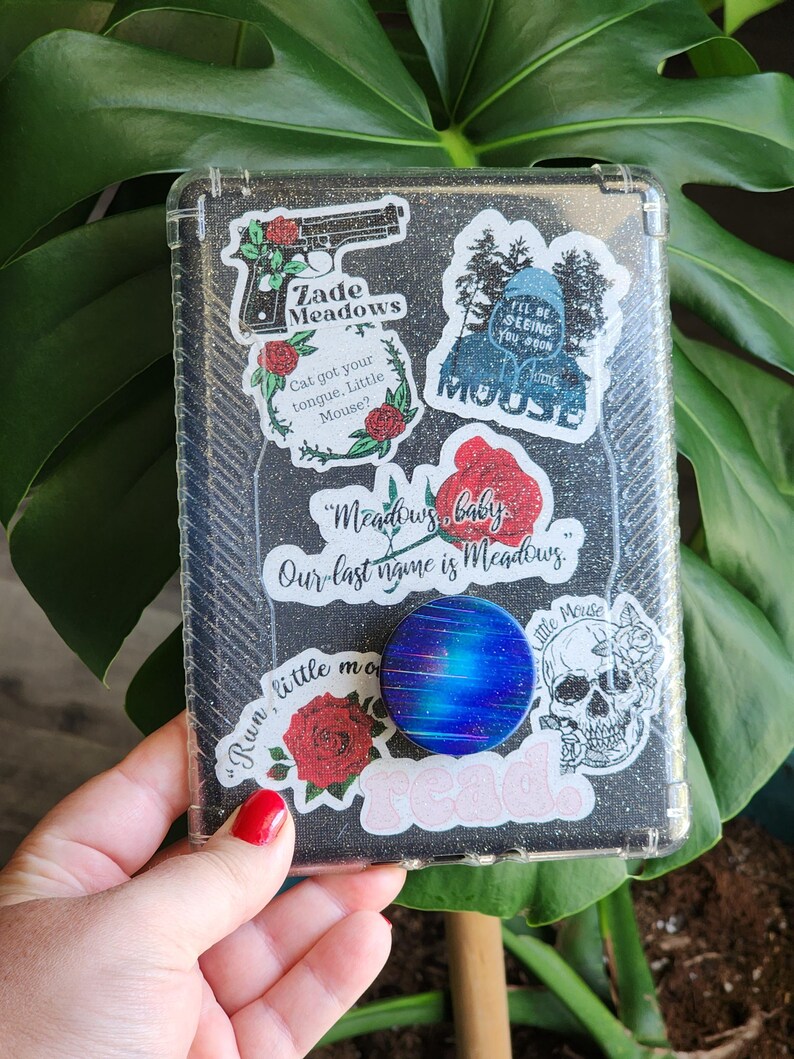 Peut inclure: Une coque de t&eacute;l&eacute;phone transparente avec un fond noir paillet&eacute;, orn&eacute;e de divers autocollants. Les autocollants incluent un pistolet avec le texte "Zade Meadows", des roses, une t&ecirc;te de mort et les mots "Meadows, baby". Un pop socket bleu et violet est attach&eacute;.