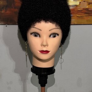 May include: A mannequin head featuring a tall, voluminous black afro hairstyle with tight curls. The mannequin has light skin, brown eyes, and red lips, and is wearing long, thin gold-toned dangling earrings with circular and linear geometric shapes.