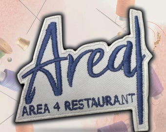 May include: White embroidered patch with blue lettering that says "Area" in a cursive font and "Area 4 Restaurant" in a smaller font.