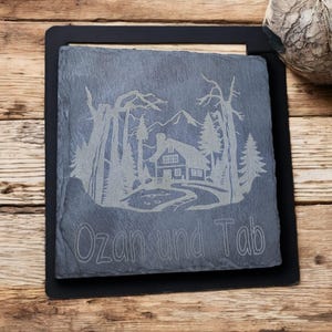 Könnte beinhalten: Ein schwarzer Rahmen aus Schiefer mit einem weißen, gravierten Bild eines Hauses in einem Wald. Der Text "Ozan und Tab" ist unter dem Bild graviert.