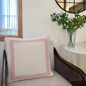 May include: A square, off-white crochet pillow with a light pink border. The pillow is placed on a light gray chair. A clear glass vase with green leaves sits on a marble table in the background.