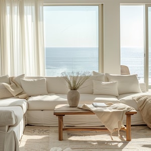 May include: A bright, airy living room with a large white sectional sofa, pillows, and a wooden coffee table. A vase with dried flowers sits on the table, and a blanket is draped over the sofa. The room has large windows overlooking the ocean.