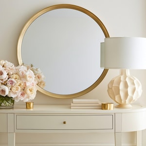 May include: A cream-colored console table with a gold-framed round mirror, a white lamp, and a bouquet of pink and cream flowers. Two small gold candle holders and a stack of books are on the table. The wall is a light cream color.