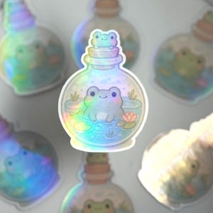 May include: A set of iridescent holographic stickers featuring frogs in glass bottles. The stickers are perfect for adding a whimsical touch to journals, laptops, water bottles, and more.