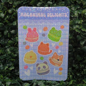 May include: A light blue sticker with the text "Macanimal Delights" and illustrations of animal-shaped macarons. The macarons are in the shape of a rabbit, frog, cat, panda, and corgi, with colorful sprinkles and details.