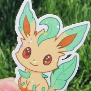 May include: A colorful sticker featuring a cartoon character with a light yellow body, green leaves, and orange accents. The character has large red eyes and a small smile. The sticker has a holographic, glittery finish, and is set against a blurred green background.