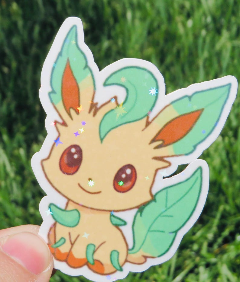 May include: A colorful sticker featuring a cartoon character with a light yellow body, green leaves, and orange accents. The character has large red eyes and a small smile. The sticker has a holographic, glittery finish, and is set against a blurred green background.