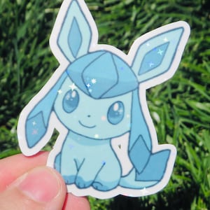 May include: A light blue sticker featuring a cartoon character with large ears and a smiling face. The sticker has a glittery, holographic effect, with star-shaped sparkles. The character is set against a background of green grass.