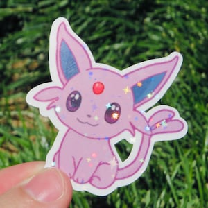 May include: A pink sticker featuring a cartoon Espeon character with blue ears, a red gem, and sparkling accents. The sticker is held against a background of green grass.