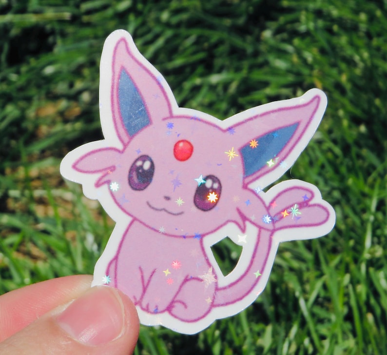 May include: A pink sticker featuring a cartoon Espeon character with blue ears, a red gem, and sparkling accents. The sticker is held against a background of green grass.