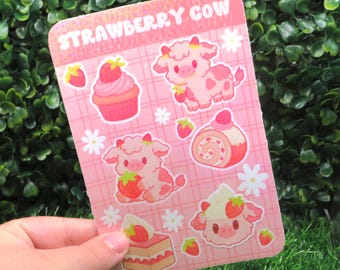 May include: A pink sticker sheet with the text "STRAWBERRY COW" features illustrations of strawberry-themed cows, cupcakes, and desserts. The sheet has a pink plaid background with white flower and strawberry accents.