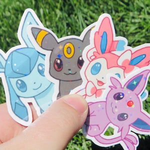May include: A hand holds four colorful stickers featuring cartoon characters. The stickers include a light blue, a gray, a pink, and a purple character. Each sticker has a white border and a glittery effect.