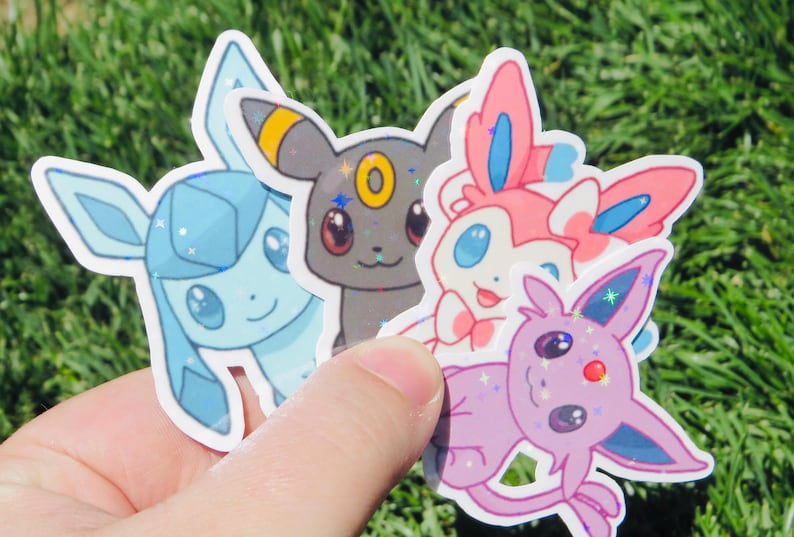 May include: A hand holds four colorful stickers featuring cartoon characters. The stickers include a light blue, a gray, a pink, and a purple character. Each sticker has a white border and a glittery effect.