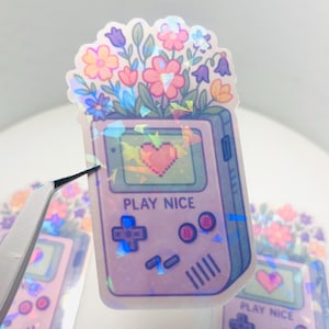 May include: A holographic sticker featuring a purple and white Game Boy with a pixelated heart on the screen. The Game Boy is surrounded by a cluster of colorful flowers and the text "PLAY NICE" is printed on the screen.