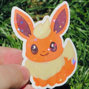 May include: A sticker featuring a cartoon Flareon character. The sticker has orange, yellow, and white coloring with glitter accents. The character has large ears, a fluffy tail, and a smiling expression. The sticker is held against a background of green grass.