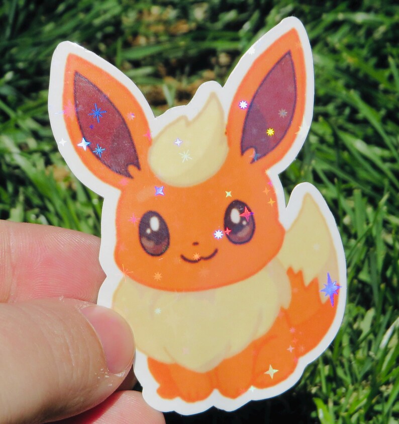 May include: A sticker featuring a cartoon Flareon character. The sticker has orange, yellow, and white coloring with glitter accents. The character has large ears, a fluffy tail, and a smiling expression. The sticker is held against a background of green grass.