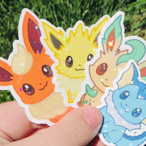 May include: A hand holding a collection of colorful stickers featuring cartoon characters. The stickers include orange, yellow, green, and blue creatures with large eyes and glitter accents. The stickers are on a green background.