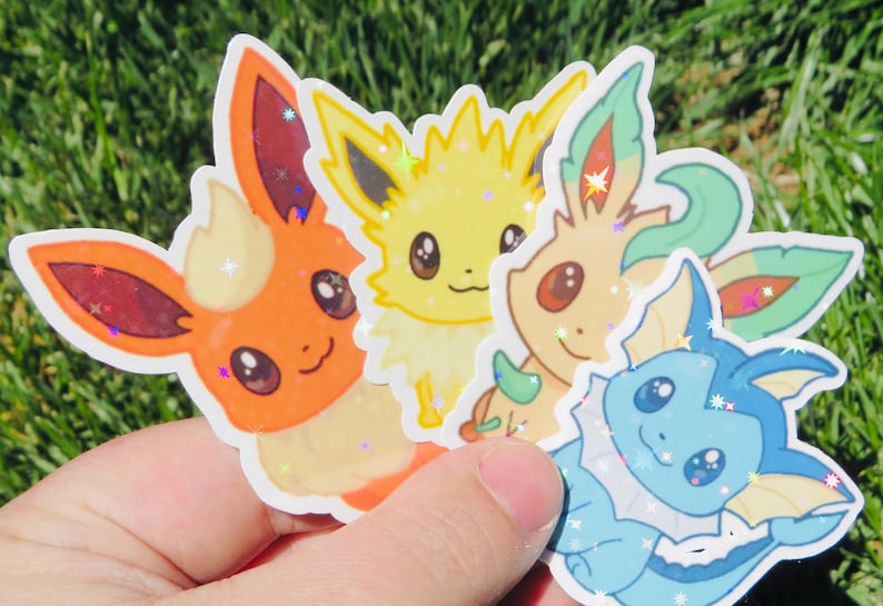 May include: A hand holding a collection of colorful stickers featuring cartoon characters. The stickers include orange, yellow, green, and blue creatures with large eyes and glitter accents. The stickers are on a green background.