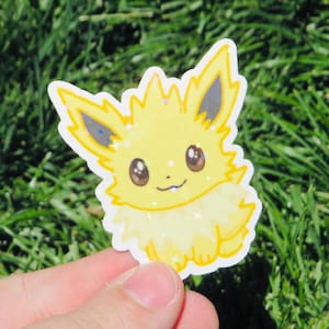 May include: A yellow sticker featuring a cartoon character with large brown eyes, pointed ears, and a fluffy tail. The sticker has a glittery effect and is held against a background of green grass.