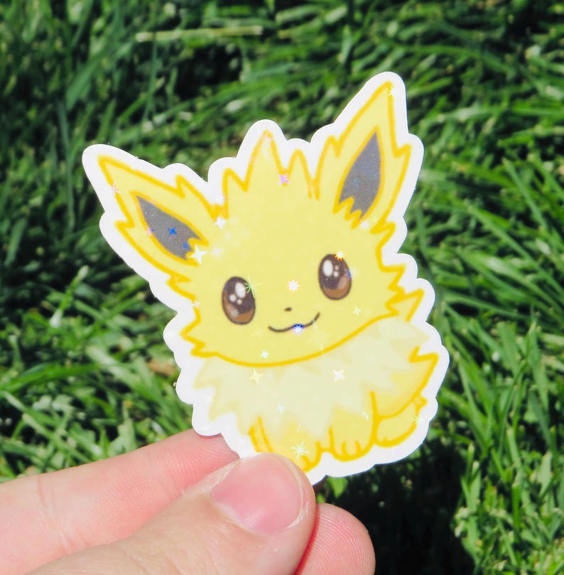 May include: A yellow sticker featuring a cartoon character with large brown eyes, pointed ears, and a fluffy tail. The sticker has a glittery effect and is held against a background of green grass.