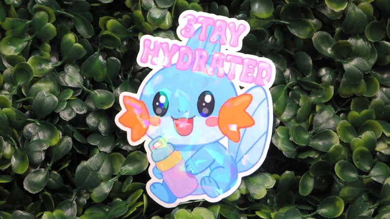 May include: A sticker featuring a blue cartoon creature holding a water bottle, with the text "STAY HYDRATED" in pink above it. The sticker has a holographic effect and is set against a background of green leaves.