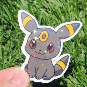 May include: A holographic sticker featuring a cartoon Umbreon character. The sticker has a gray body with yellow accents on its ears and tail. The eyes are red, and the sticker has a sparkly, iridescent finish. The character is sitting down.
