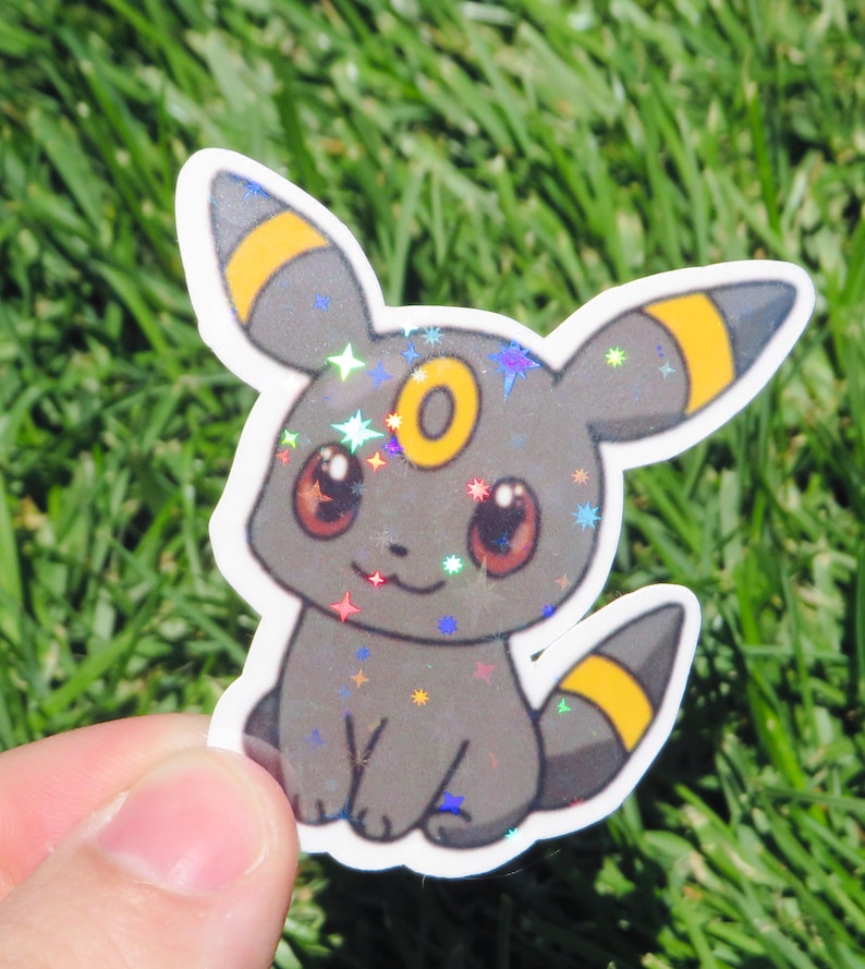 May include: A holographic sticker featuring a cartoon Umbreon character. The sticker has a gray body with yellow accents on its ears and tail. The eyes are red, and the sticker has a sparkly, iridescent finish. The character is sitting down.