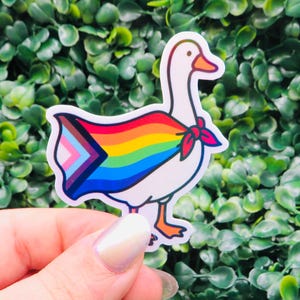 May include: A white goose sticker with an orange beak and feet, wearing a rainbow cape with the Progress Pride flag design. The sticker has a red bow tie and is set against a green leafy background.