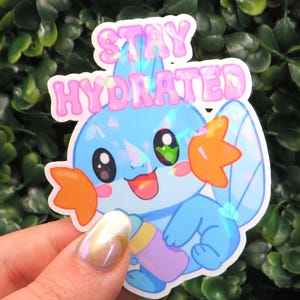 May include: A holographic sticker featuring a cartoon character with blue and orange accents, holding a bottle. The sticker has the text "STAY HYDRATED" in pink bubble letters. The sticker is held in front of a green leafy background.