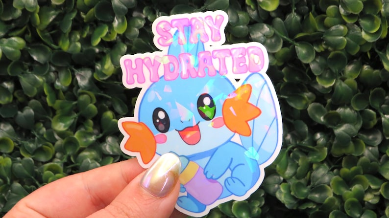 May include: A holographic sticker featuring a cartoon character with blue and orange accents, holding a bottle. The sticker has the text "STAY HYDRATED" in pink bubble letters. The sticker is held in front of a green leafy background.