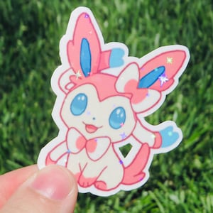 May include: A die-cut sticker featuring a cartoon character with pink, white, and blue accents. The character has large blue eyes, a pink bow, and glitter details. The sticker is held against a background of green grass.