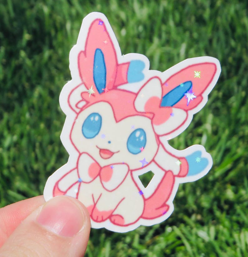 May include: A die-cut sticker featuring a cartoon character with pink, white, and blue accents. The character has large blue eyes, a pink bow, and glitter details. The sticker is held against a background of green grass.