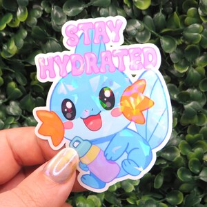 May include: A holographic sticker featuring a cartoon character with blue, orange, and pink accents, holding a water bottle. The sticker has the words "STAY HYDRATED" in pink bubble letters.