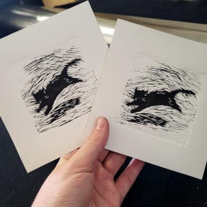 K&ouml;nnte beinhalten: Zwei Linolschnittdrucke eines rennenden Katers in schwarzer Tinte auf wei&szlig;em Papier. Die Silhouette der Katze ist mit strukturierten Linien detailliert, vor einem Hintergrund aus &auml;hnlichen Linien, die Bewegung suggerieren. Die Drucke werden in einer Hand gehalten.