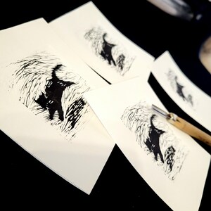 K&ouml;nnte beinhalten: Eine Sammlung von Linolschnitten mit einer schwarzen Katzen-Silhouette in einer strukturierten, abstrakten Landschaft. Die Drucke befinden sich auf wei&szlig;em Papier, mit einem Schnitzwerkzeug im Hintergrund. Das Kunstwerk wird auf einer dunklen Oberfl&auml;che pr&auml;sentiert.