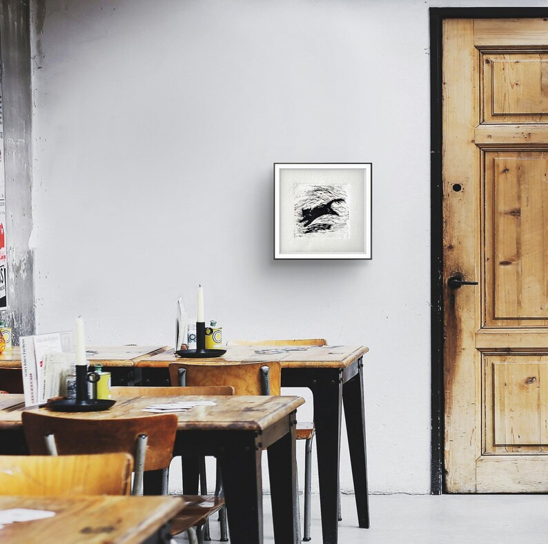 K&ouml;nnte beinhalten: Ein gerahmter Schwarz-Wei&szlig;-Druck eines springenden Tieres, m&ouml;glicherweise einer Katze oder eines Hundes, vor einem strukturierten Hintergrund. Das Kunstwerk wird an einer wei&szlig;en Wand in einem Raum mit Holztischen und einer Holzt&uuml;r ausgestellt.