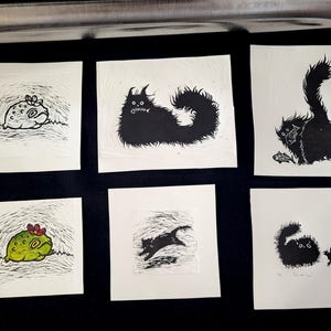 K&ouml;nnte beinhalten: Eine Sammlung von sechs quadratischen Linolschnitten mit skurrilen Schwarz-Wei&szlig;-Katzenillustrationen. Ein Druck zeigt einen Frosch mit einer Blume, ein anderer eine Katze mit einem Fisch. Die Drucke sind auf wei&szlig;em Papier.